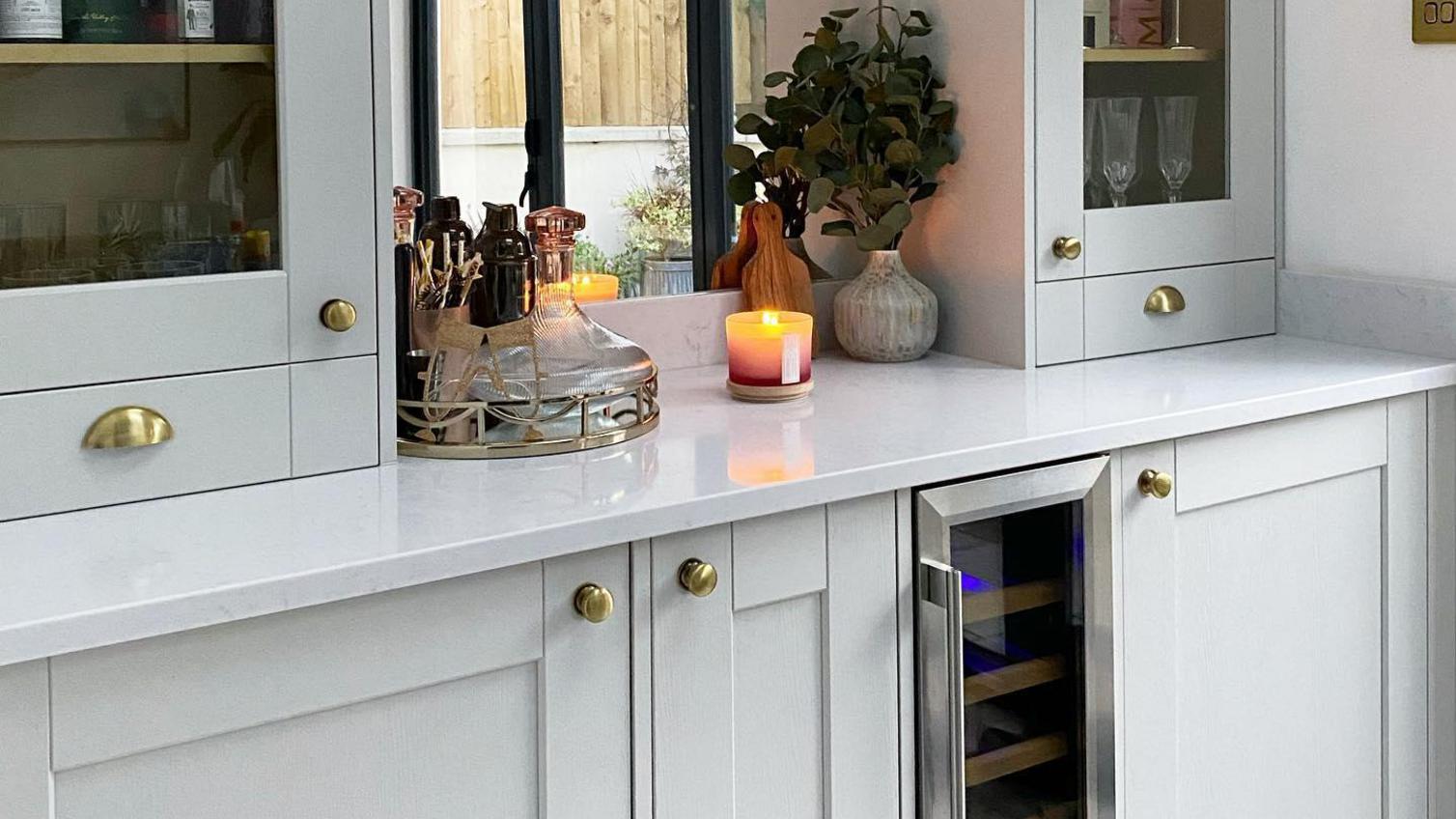 Light grey shaker bar idea with brass cup and knob handles, glass cupboard doors, a wine fridge and quartz worktop.