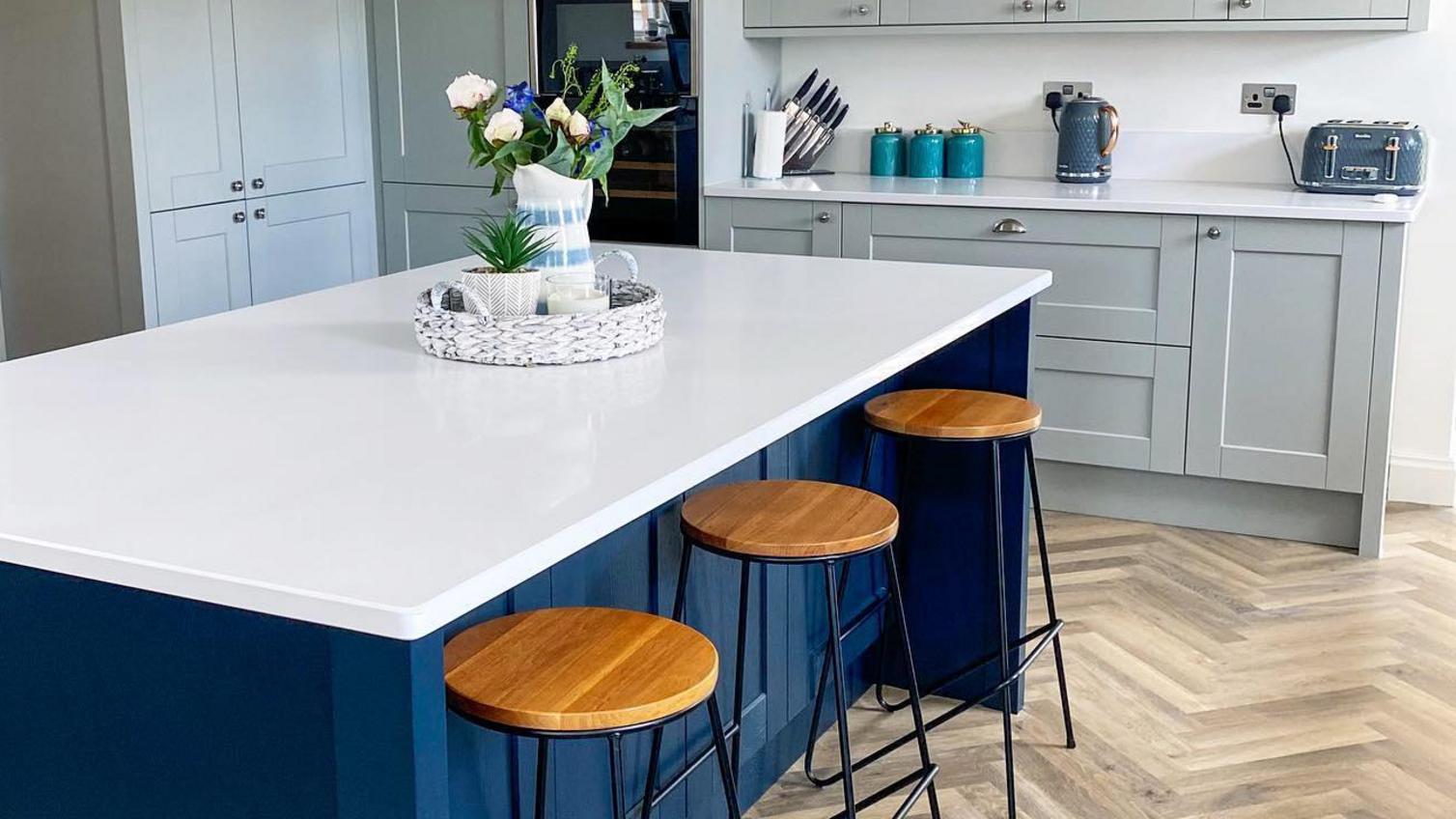 Navy kitchen island with dove-grey cabinets, a white worktop, oak bar stools, shaker doors and chevron floors.