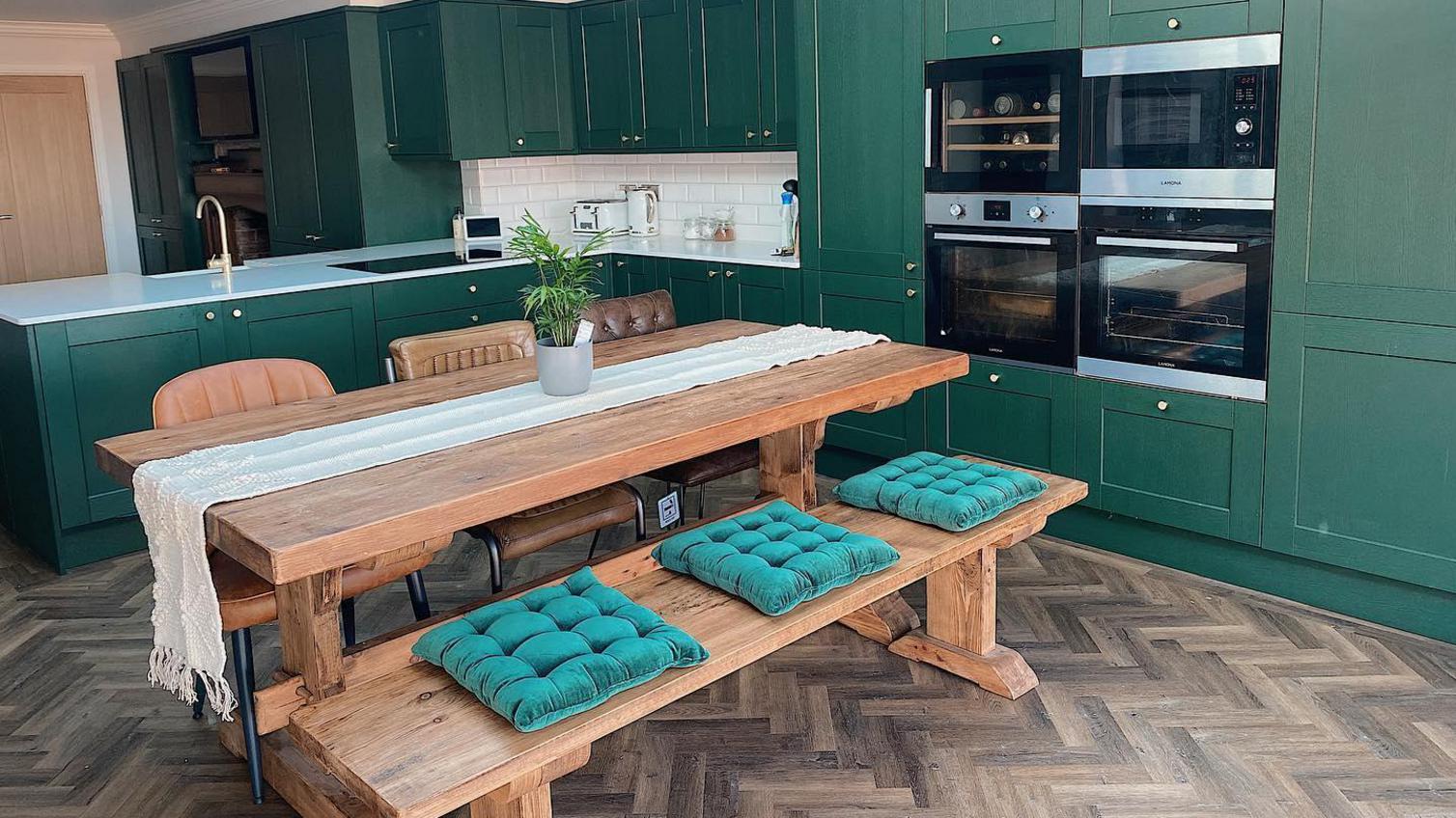 Dark green shaker kitchen ideas with gold knob handles, built in cooking devices, and an oak bench table with green cushions.
