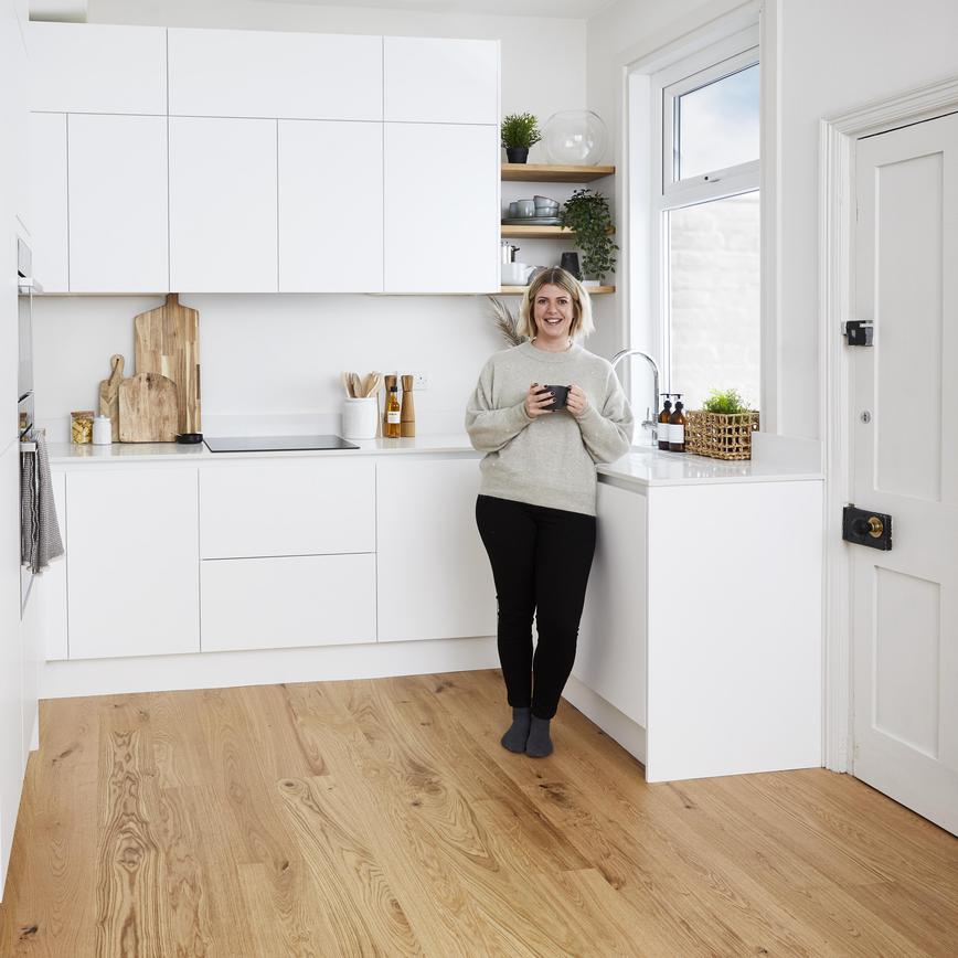 Woman standing in an u-shaped modern white kitchen. Has wooden flooring, white quartz worktops, and tower units.
