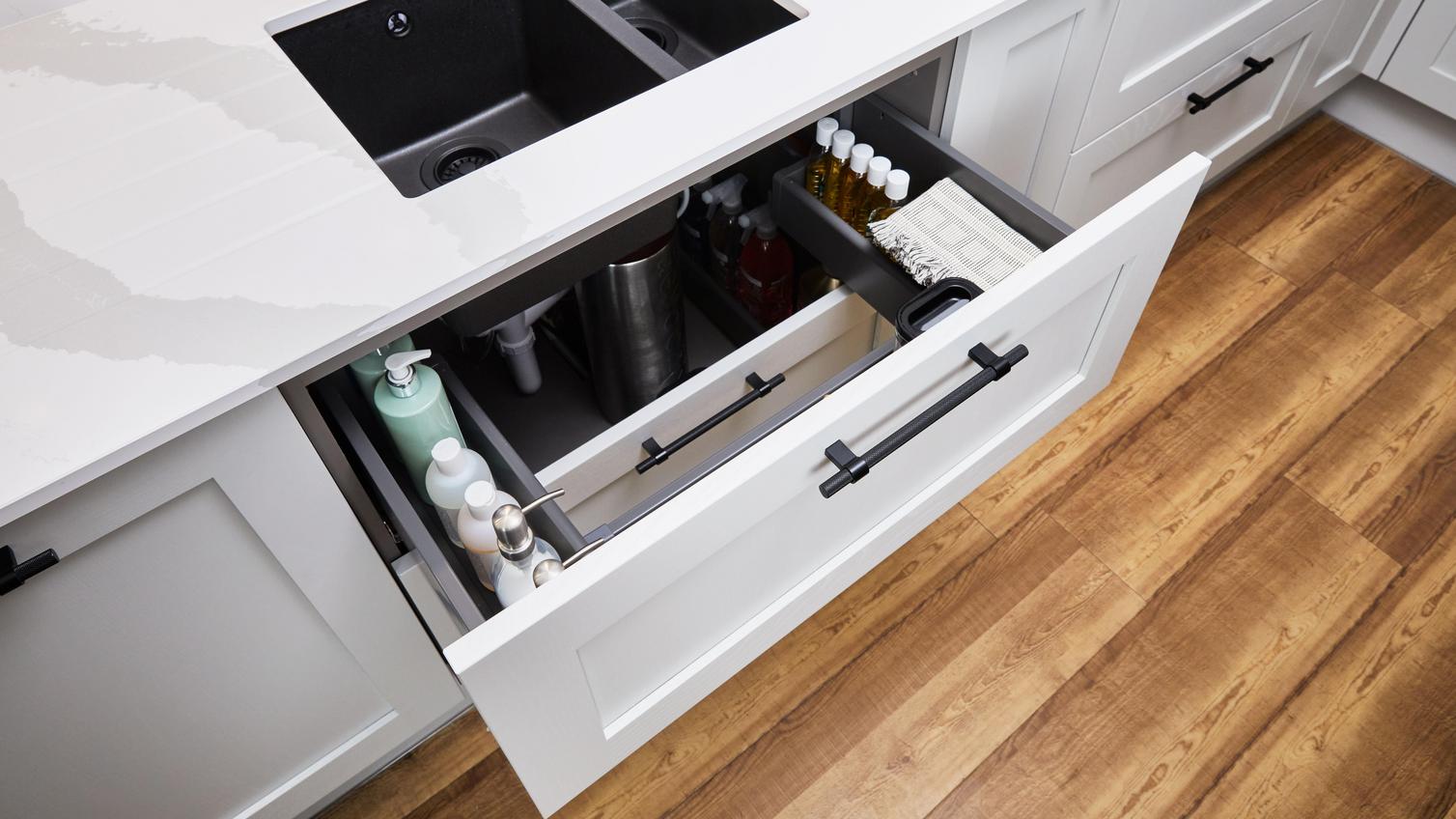 Under-sink drawer filled with cleaning products beneath a black, 1.5-bowl sink which is inset into a white quartz worktop.