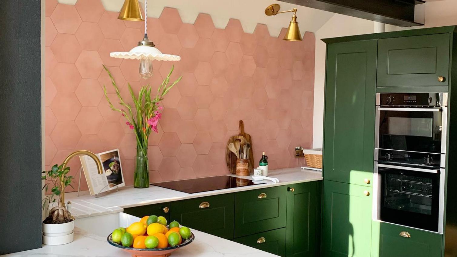 Green shaker style kitchen with a breakfast bar, featuring pink hexagon tiled wall, white marble worktop and gold handles.