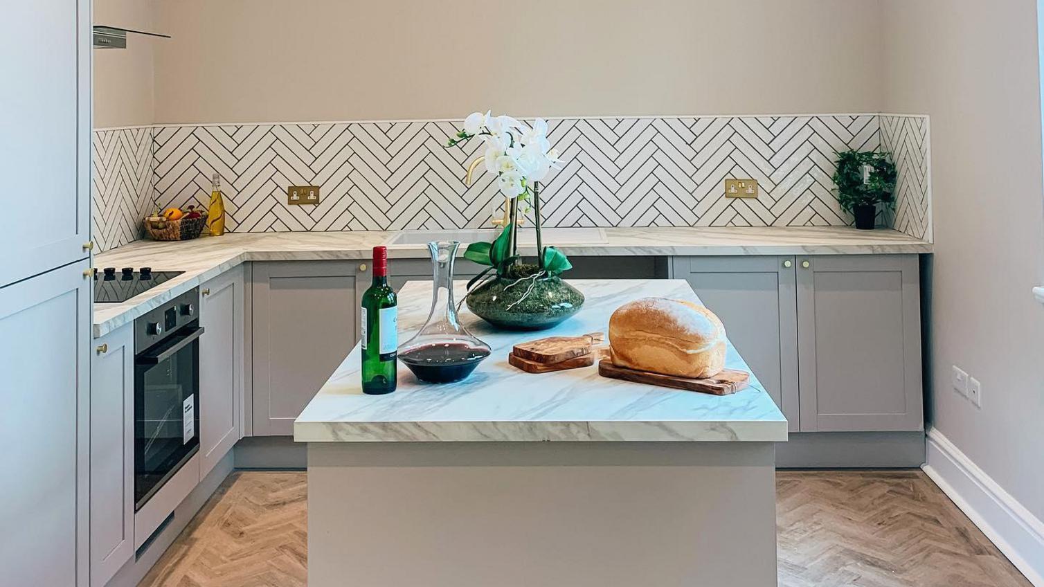 Grey shaker style kitchen with light oak chevron flooring, white marble worktops and white chevron tiles.