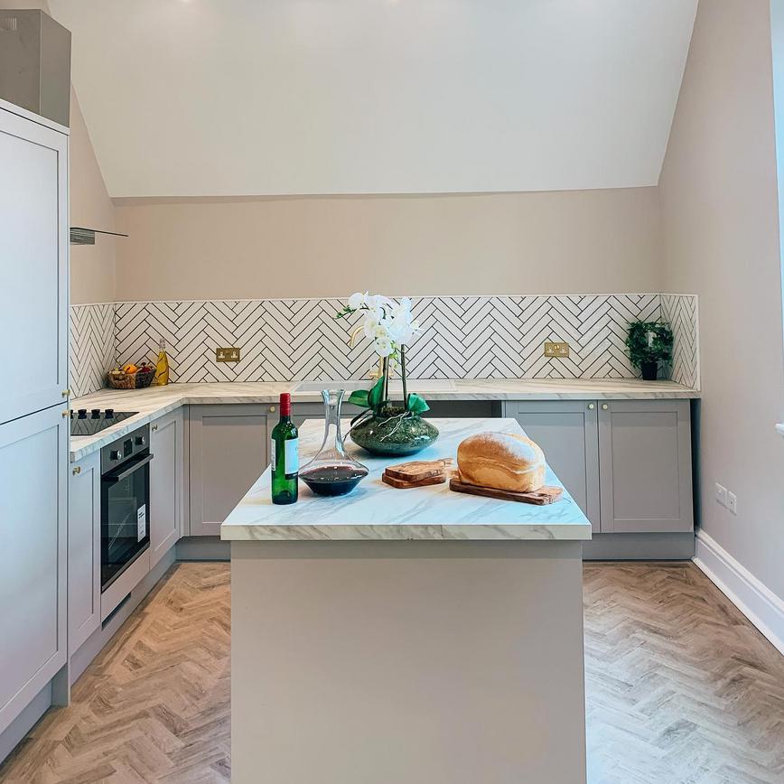 Grey shaker style kitchen with light oak chevron flooring, white marble worktops and white chevron tiles.