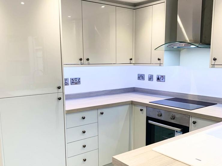 White slab kitchen design with gloss cupboards and black knob handles. Showing light oak-effect worktops and flooring.