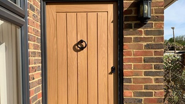 Oak panelled door with five long vertical panels, a black frame and matching black circle knocker, letterbox and handle.