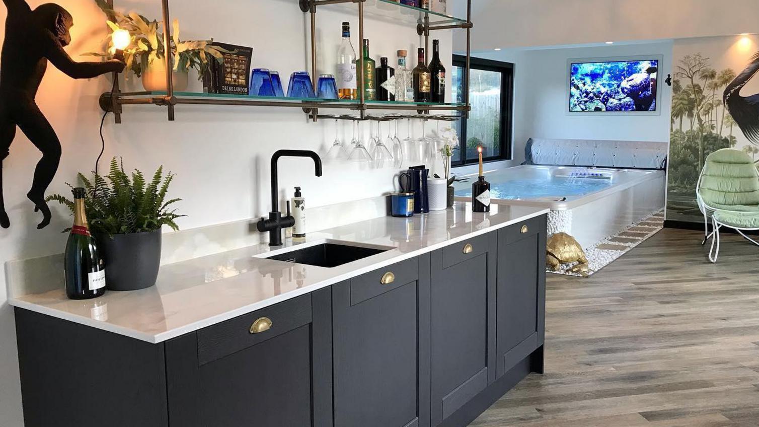Home bar idea with charcoal shaker cupboard doors, a white quarts worktop, black tap, and open shelving with bottles.