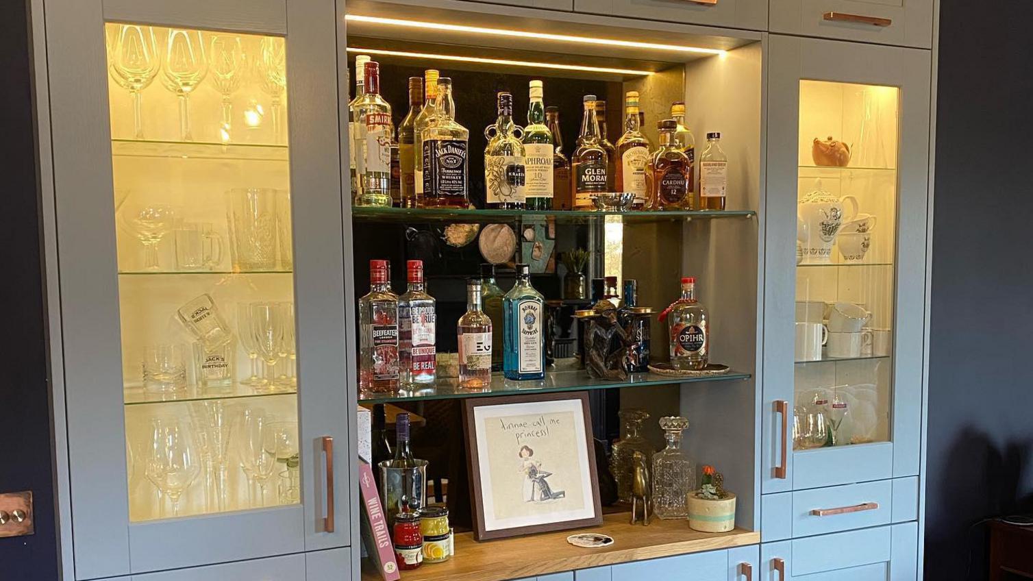 Home bar idea with light-grey shaker cupboards, two glass doors next to open shelving showing crockery and drink bottles.