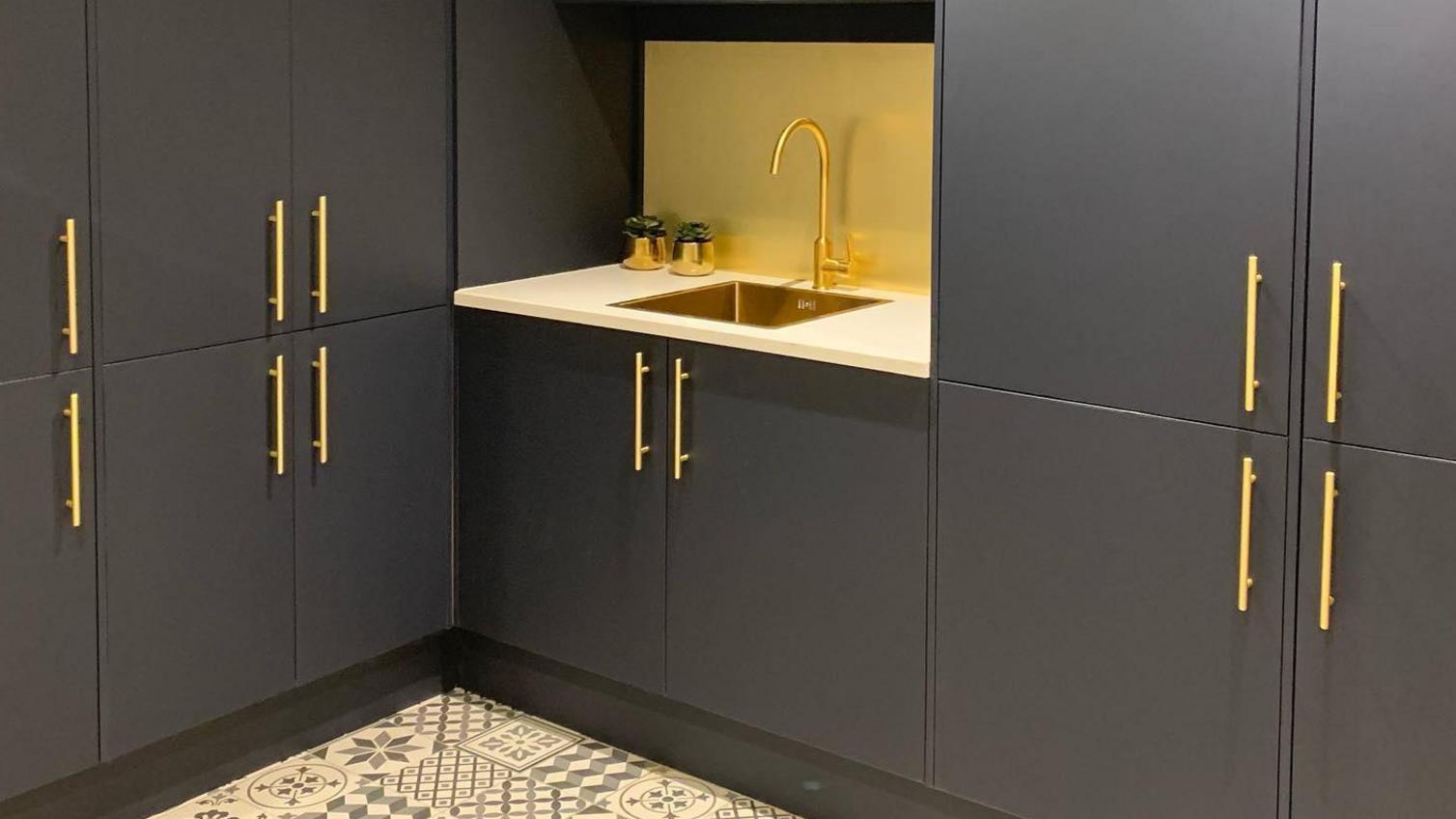 Utility room idea with banks of charcoal slab cupboards, including tower units, gold bar handles and a gold tap and sink.