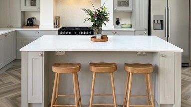 Light grey kitchen island layout with a white worktop, chrome cup handles, shaker doors, and oak stools.