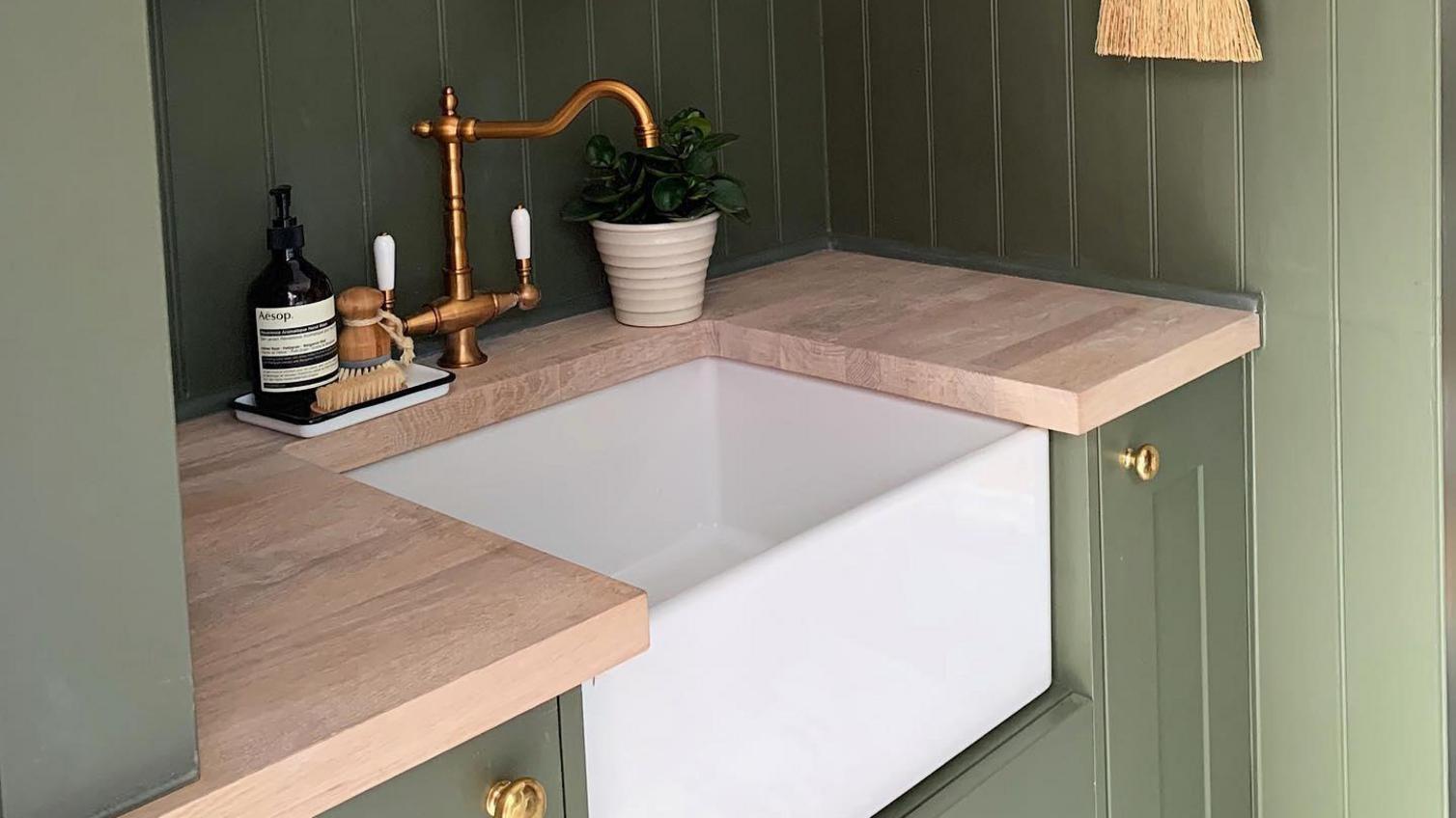Green panelled sink area with a ceramic belfast sink, light oak worktop, hanging rod with accessories and a copper tap.