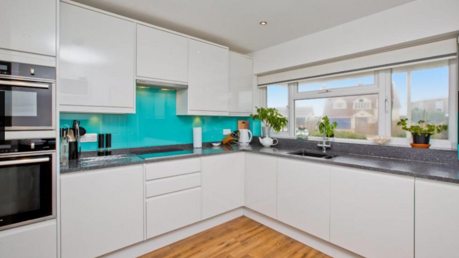 White integrated handle gloss kitchen in an l-shaped layout, with grey laminate worktops, and an aqua backsplash.