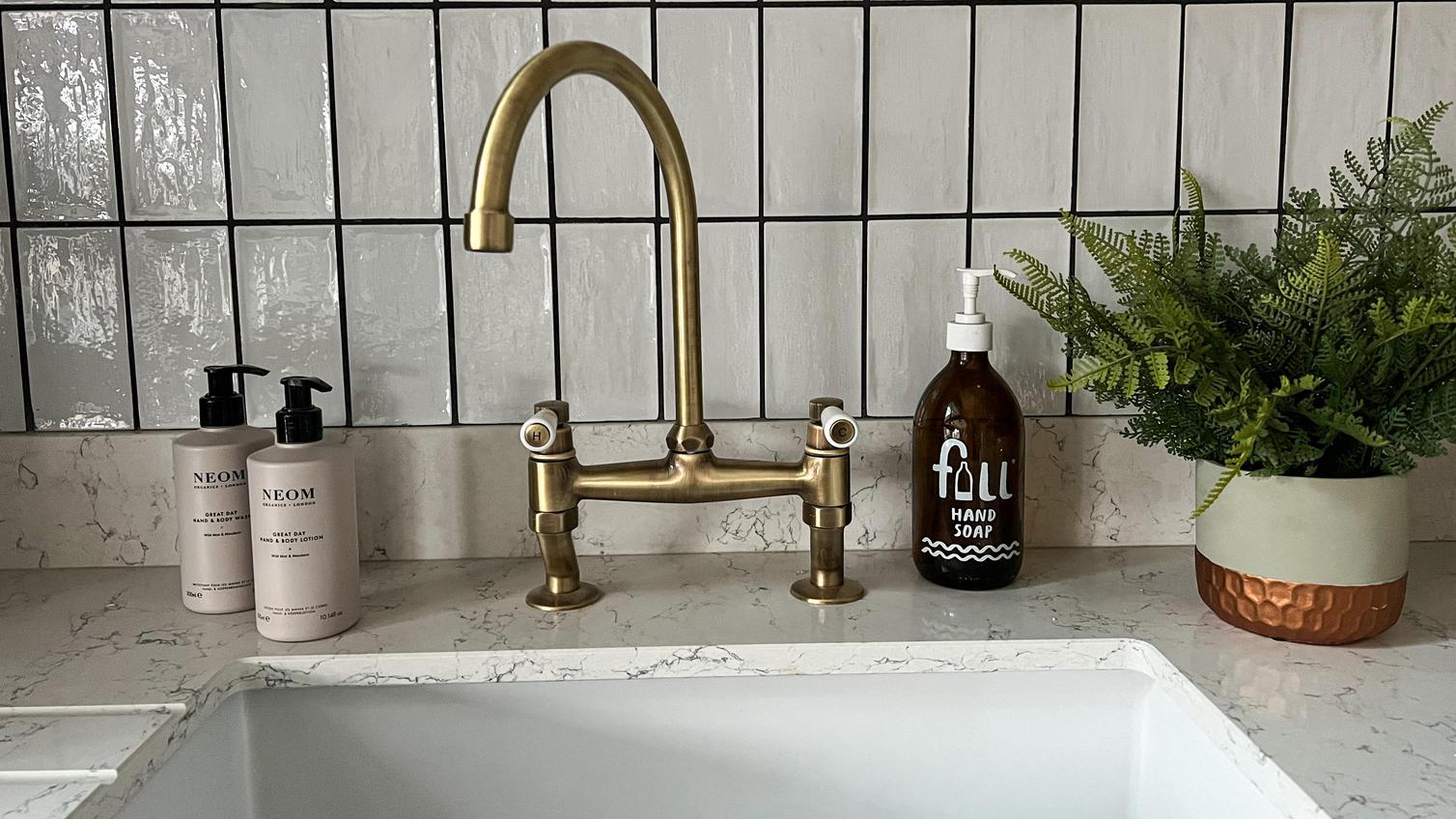 Antique brass swan neck tap with two handles, textured white subway tiles, a white quartz worktop and hand soaps.