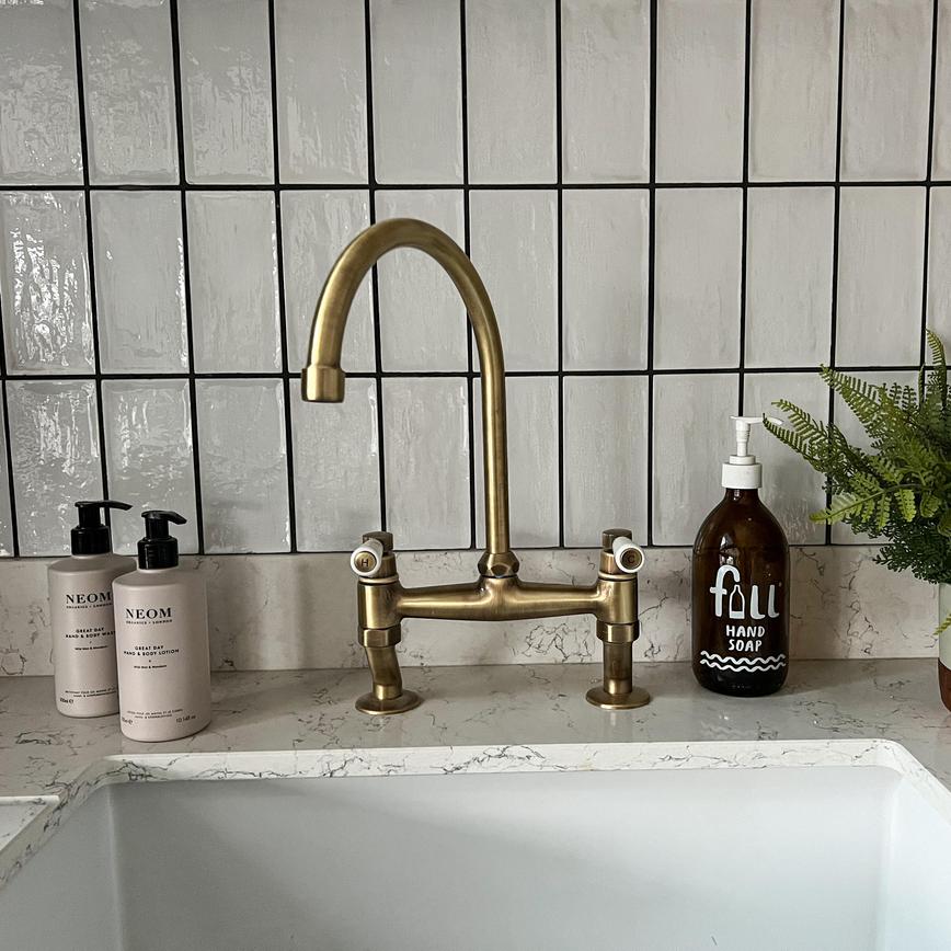 Antique brass swan neck tap with two handles, textured white subway tiles, a white quartz worktop and hand soaps.