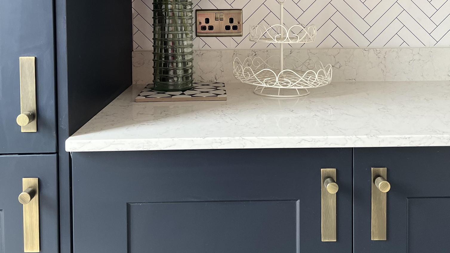 A luxe utility room idea with navy shaker units, white quartz worktops, brushed brass handles and backplates.