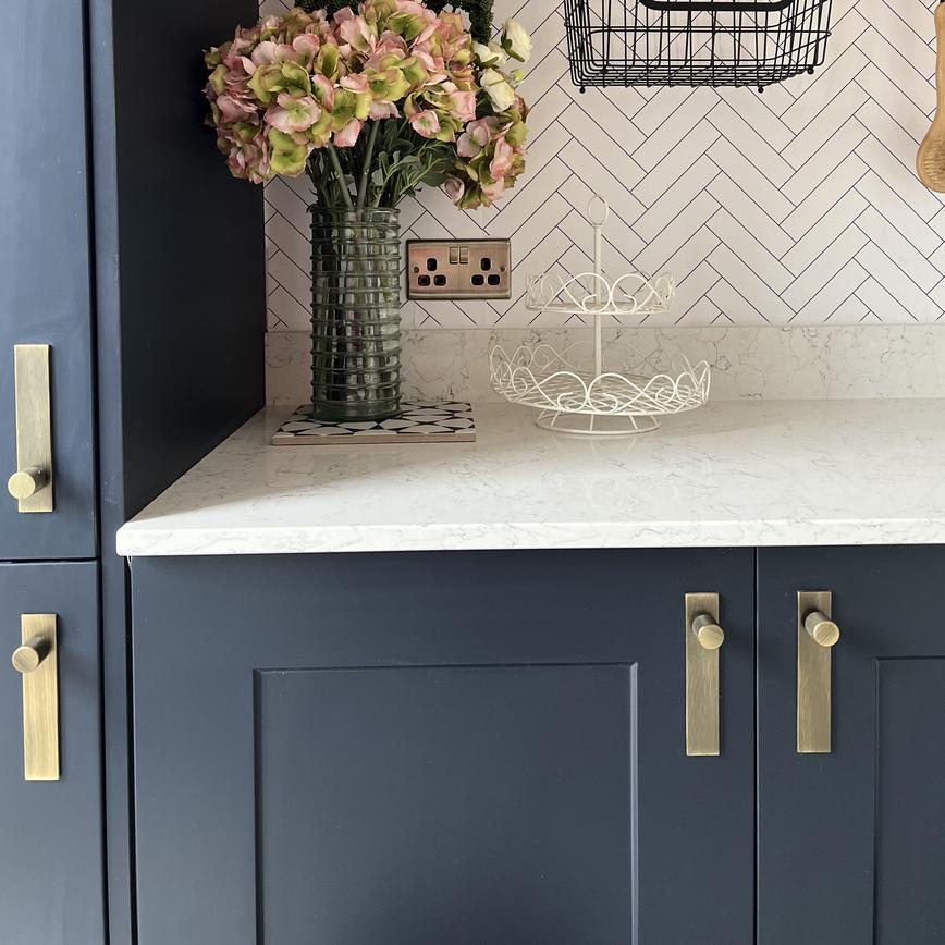 A luxe utility room idea with navy shaker units, white quartz worktops, brushed brass handles and backplates.