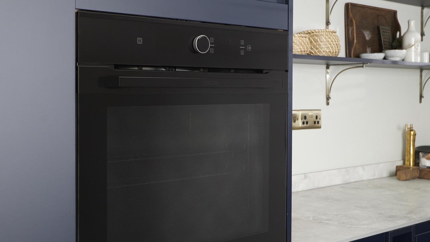 An integrated, black Lamona oven which is built into an appliance cabinet at eye-level. It is alongside navy kitchen doors.
