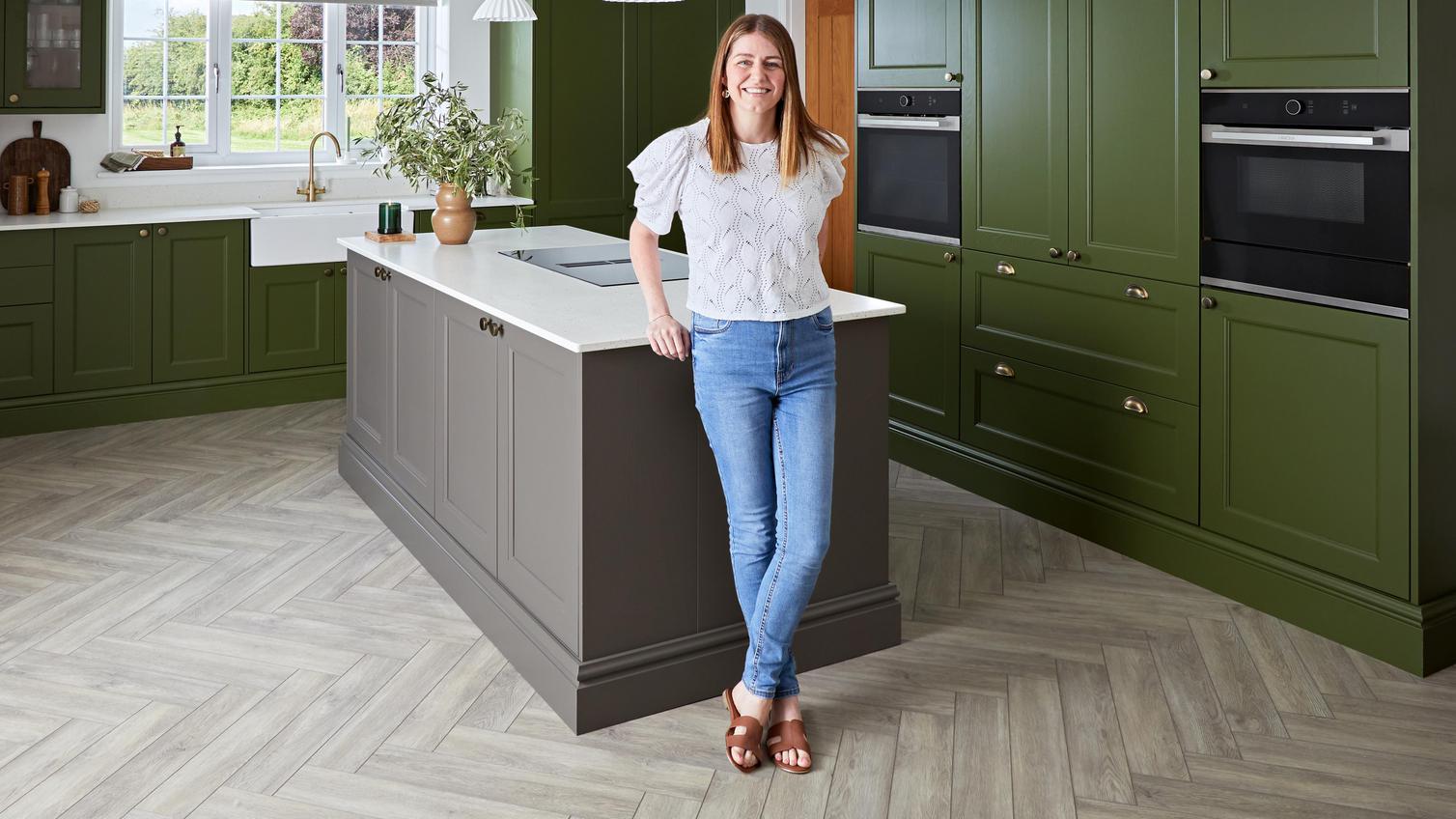 Interior influencer Kate, standing in her green and brown kitchen. It has a kitchen island as in an l-shaped layout.