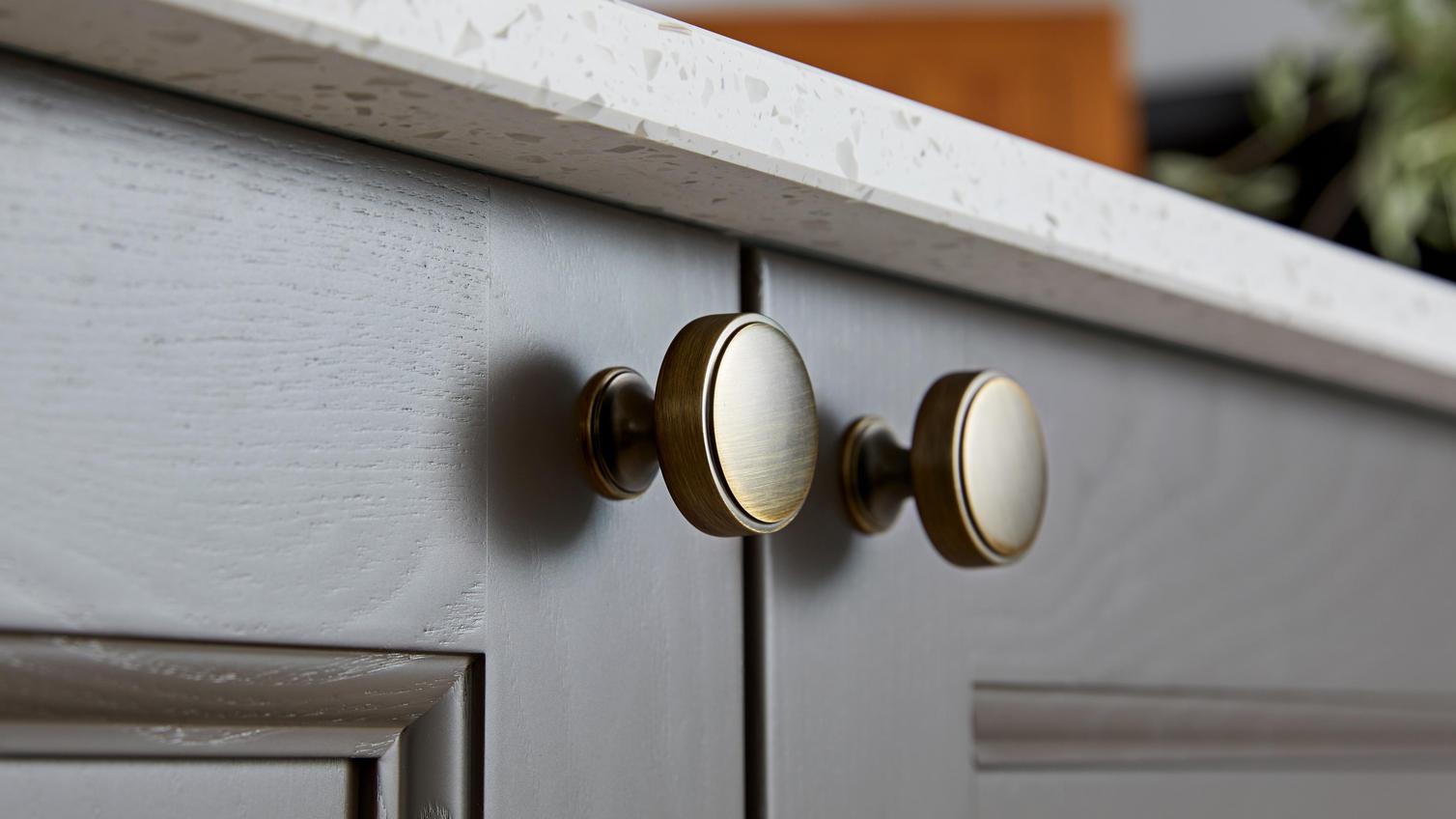 Antique brass cabinet door knobs on a brown shaker kitchen door. Above the cabinet is a white acrylic worktop.