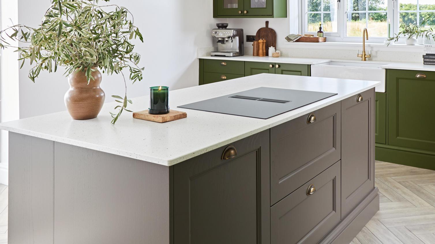 A green kitchen with a brown shaker kitchen island. It has a built-in hob, white worktop, and brass cup handles.
