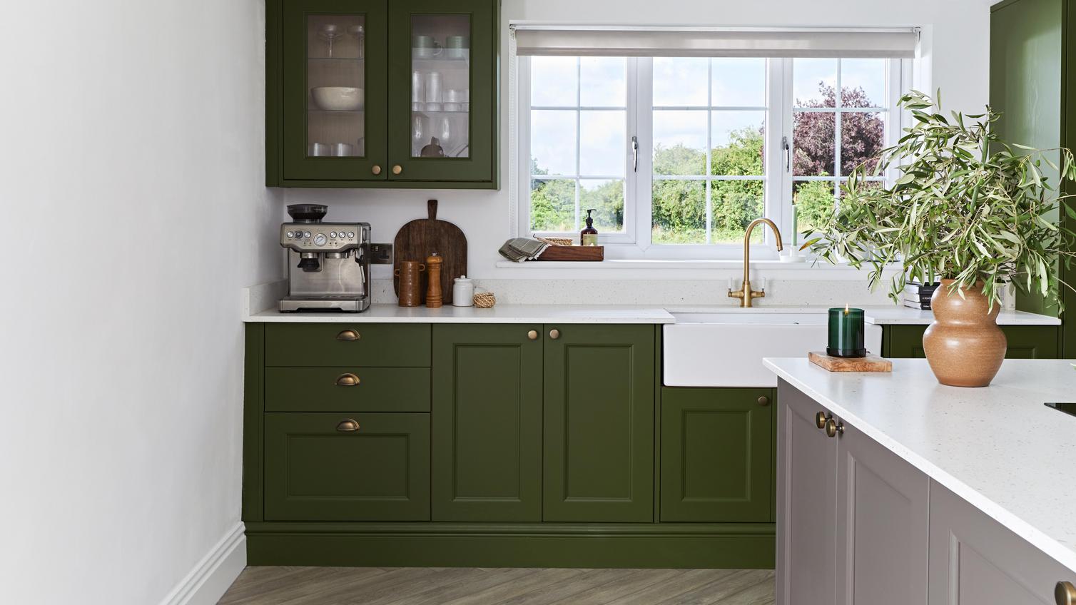 A green shaker kitchen in an olive tone. It has brass accessories, glass wall cabinets, and white worktops.