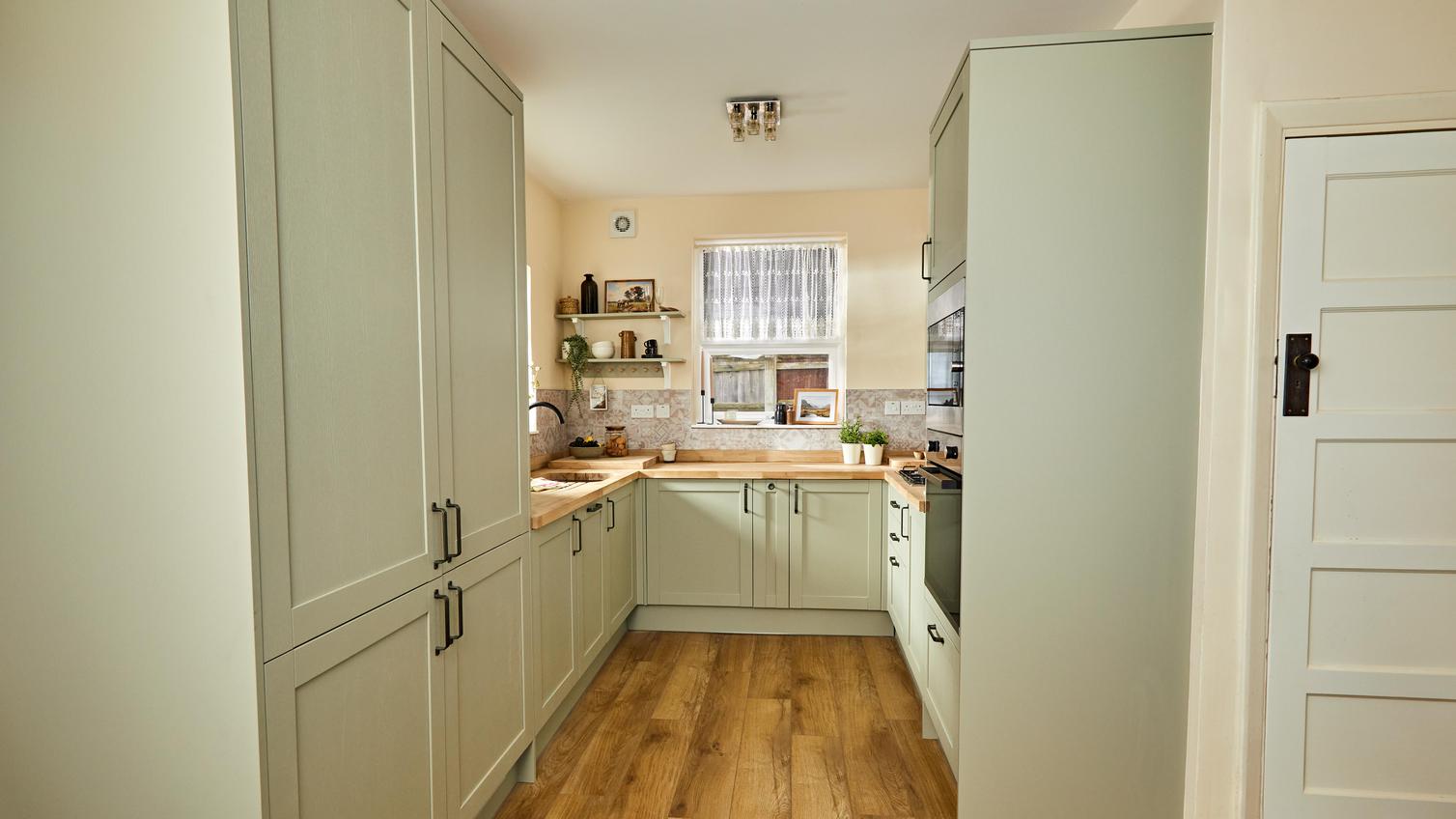 A U-shaped kitchen layout featuring sage-green cupboard doors in a shaker design, black handles, and timber worktops.