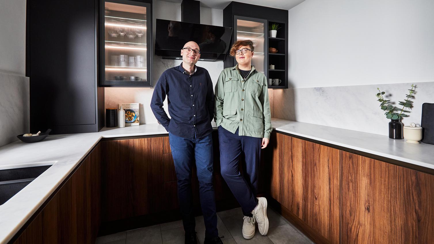Male couple standing in a modern kitchen with black and walnut slab cupboard doors. Includes white worktops and grey floors.
