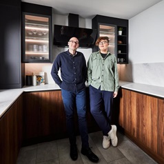 Male couple standing in a modern kitchen with black and walnut slab cupboard doors. Includes white worktops and grey floors.