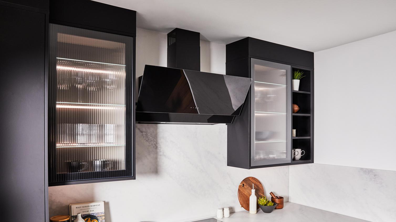 Fluted glass wall units on either side of a black, angled chimney extractor in a kitchen. Contains white backboards and worktops.