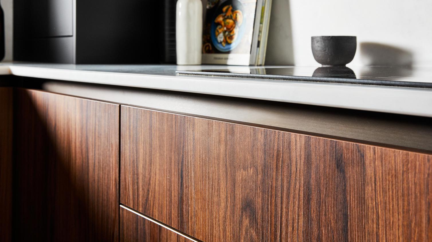 Handleless units featuring a black, metal profile along the top, walnut cupboard doors in a slab style, and white worktops.