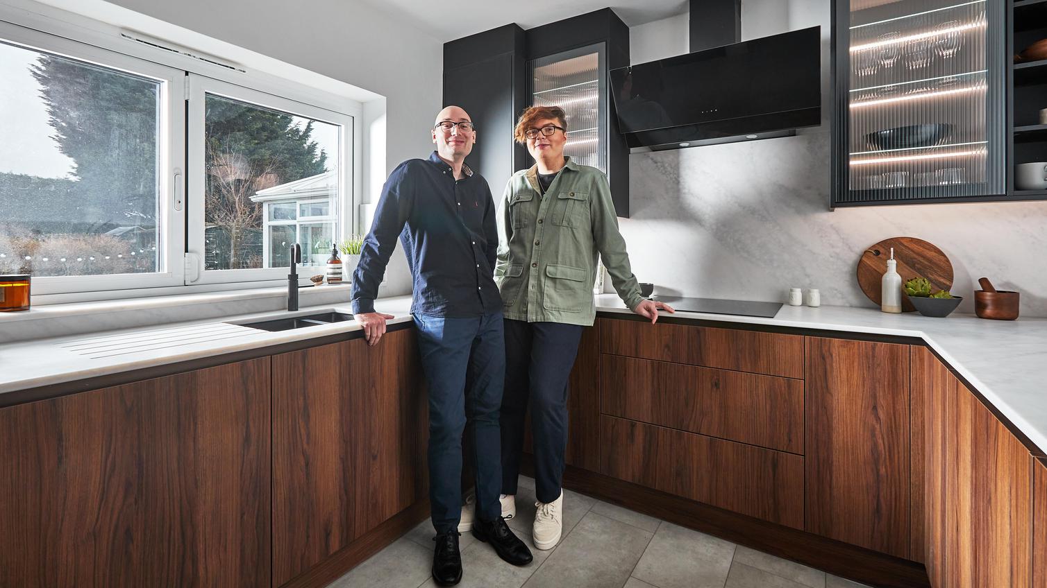 Josh and Dan standing in their kitchen which contains black wall cupboards with fluted glass doors and handleless walnut units.