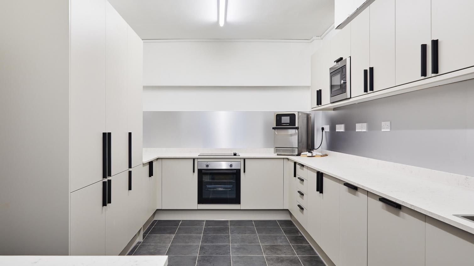 Jill Scott Cafe Kitchen Makeover with slab cupboards in a sandstone colour. Has black handles, white worktops, and grey floors.