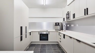 Jill Scott Cafe Kitchen Makeover with slab cupboards in a sandstone colour. Has black handles, white worktops, and grey floors.
