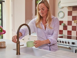 Lucy stood at the Belfast sink using the Quooker tap. It is finished in brass, and there are pink cabinets in the background