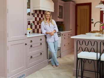 Lucy stood in her pink kitchen, leaning against the wall cabinets. There is a white worktop and a range cooker is behind her.