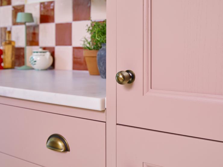 A pink shaker kitchen door and drawer styled with brass d-shaped cup handles. Behind is a white worktop and tiled splashback