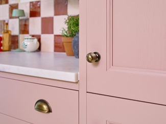 A pink shaker kitchen door and drawer styled with brass d-shaped cup handles. Behind is a white worktop and tiled splashback