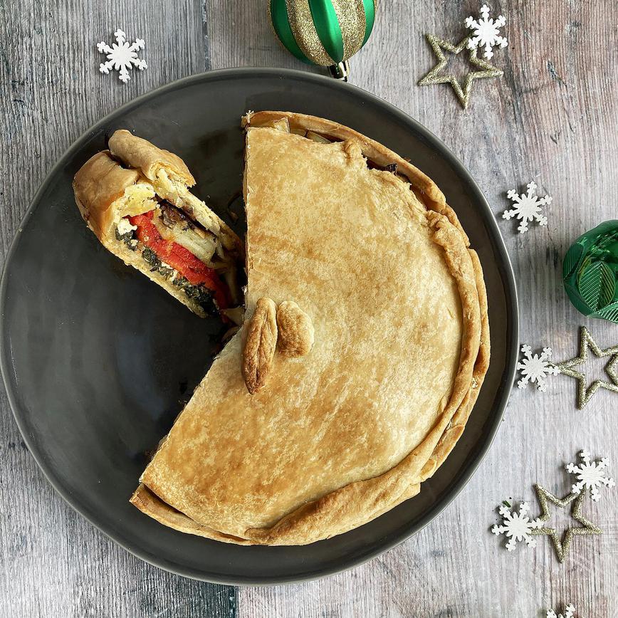 Vegan pie recipe showing a cooked pastry pie with layers of roasted vegetables on a black plate, with green baubles.