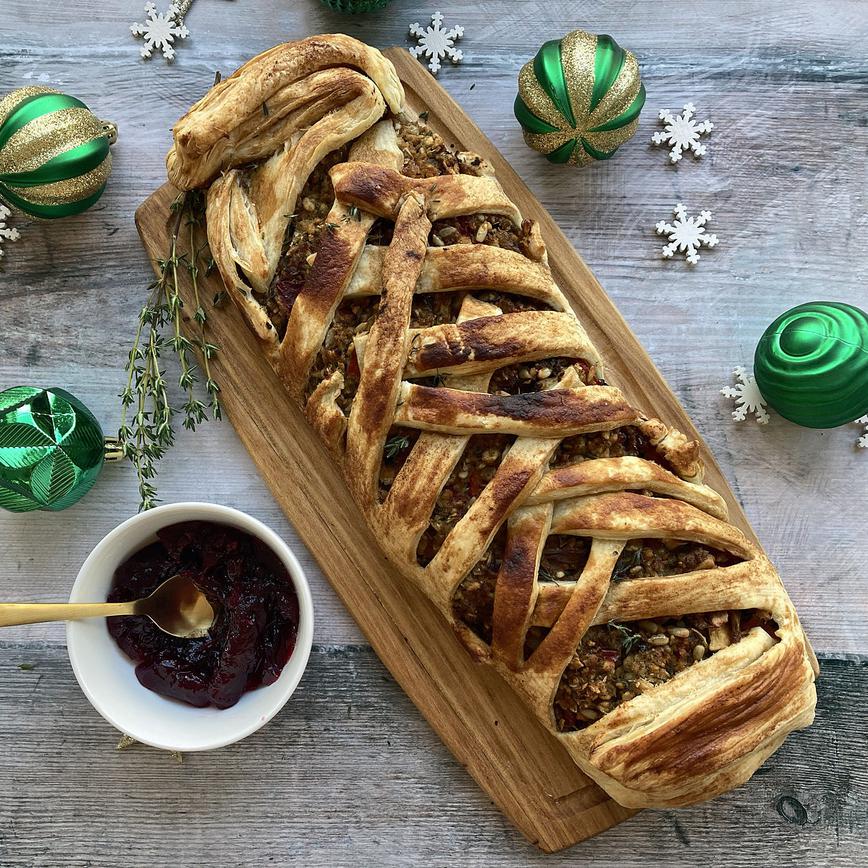 Sausage roll recipe showing a sausage and pastry plait on a wooden plank, with a dipping sauce and green baubles.