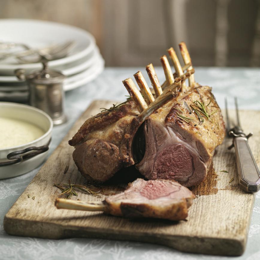Rack of lamb on a wooden cutting board, showing one side cut open and placed on the board, with a carving knife and plates.