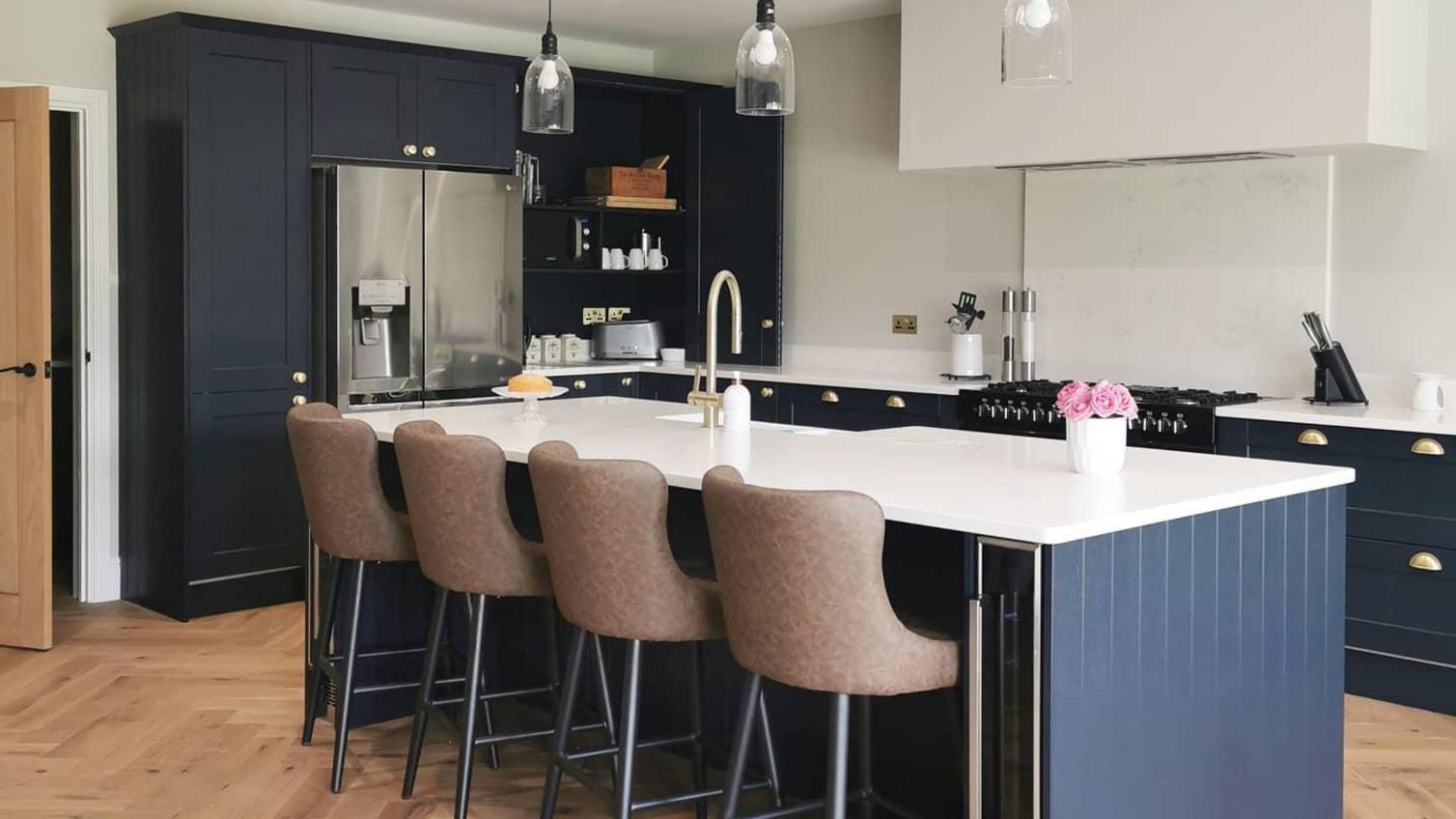 Navy blue shaker kitchen with an island layout, featuring oak chevron flooring, pendant lights and a white quarts worktop.