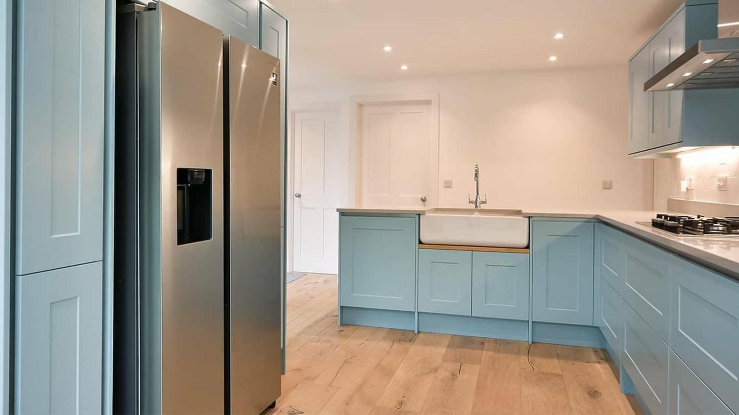 A blue kitchen idea using paintable shaker doors. Features a white, double bowl ceramic sink, wooden worktops, and floors.