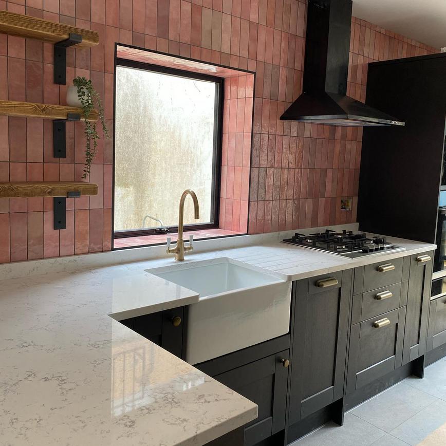 Black shaker kitchen design with a white marble worktop, a white butler sink, gold tap, and pink tiling on the walls.