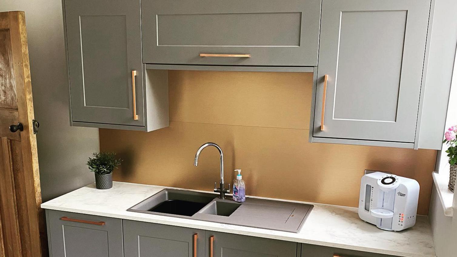 Grey shaker kitchen with brass bar handles, a white quartz worktop, gold shiny backsplash and a silver tap.