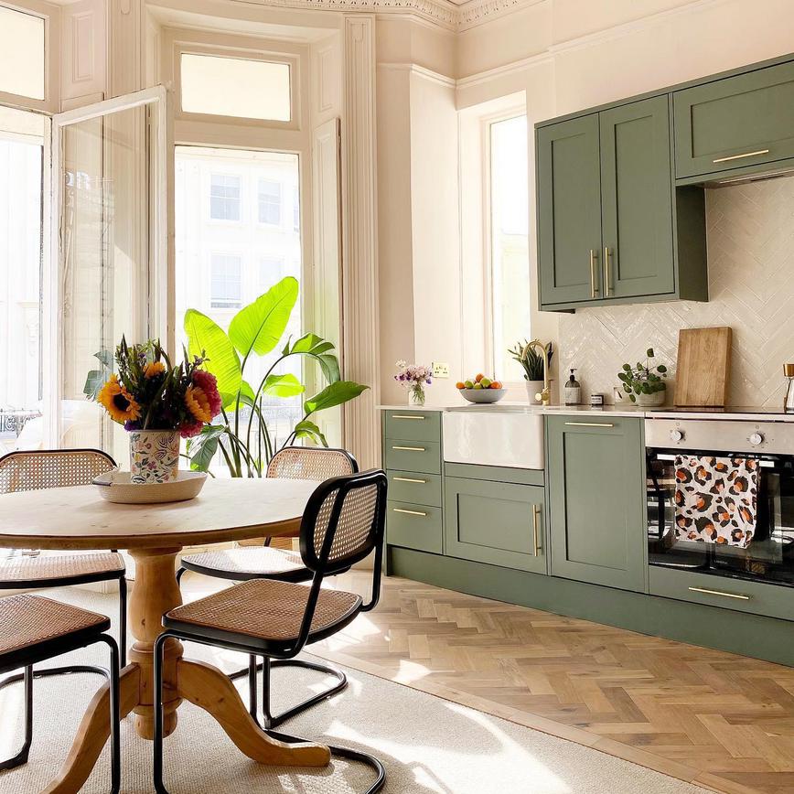 Paintable kitchen design in Victorian room with geen shaker cupboards, chevron flooring, a round table, and open windows.