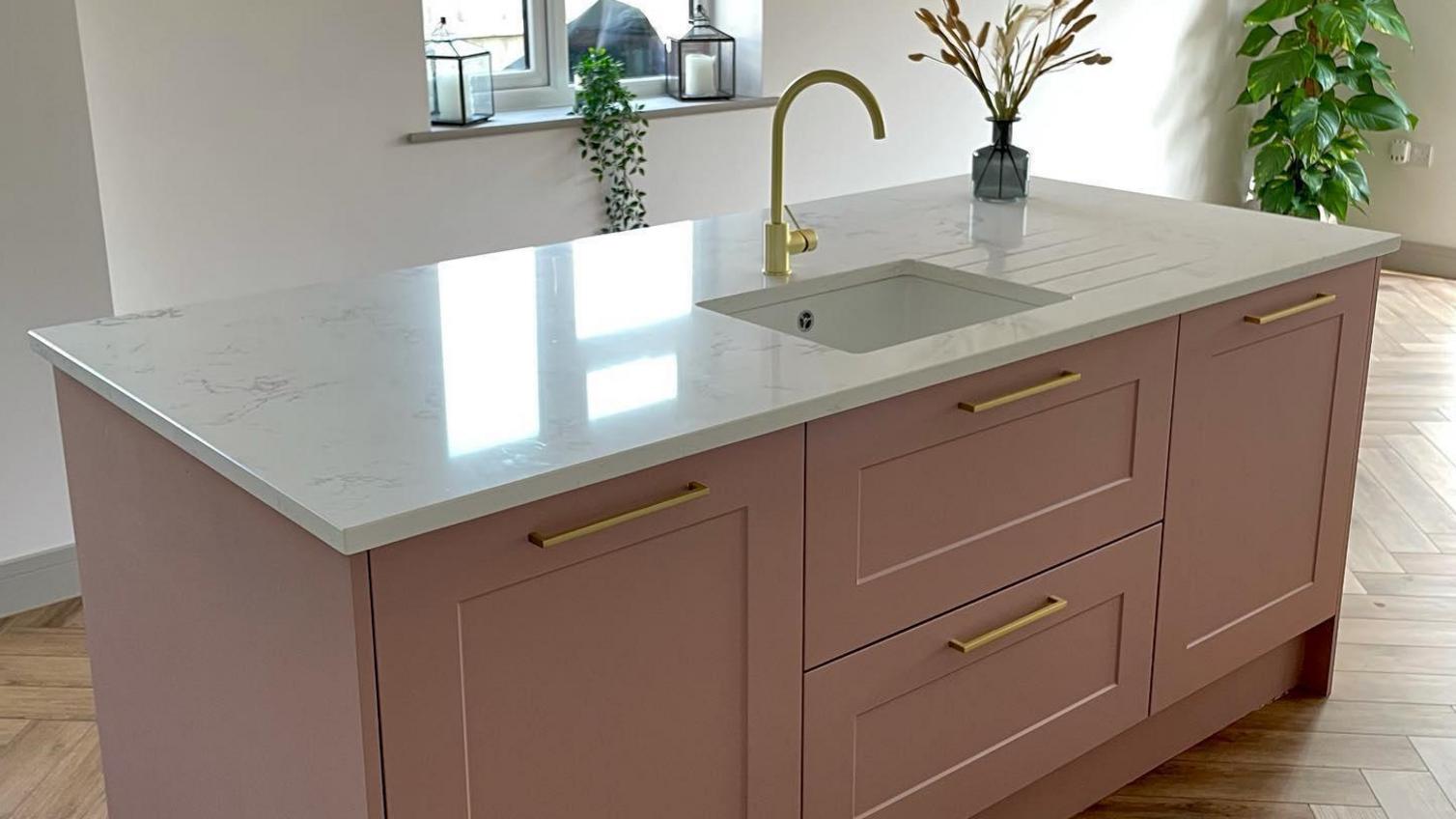 Pink kitchen island with a white quartz worktop, chevron oak flooring, gold bar handles and a gold tap.