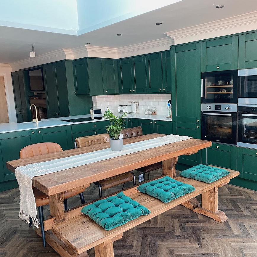 Dark green shaker kitchen ideas with gold knob handles, built in cooking devices, and an oak bench table with green cushions.