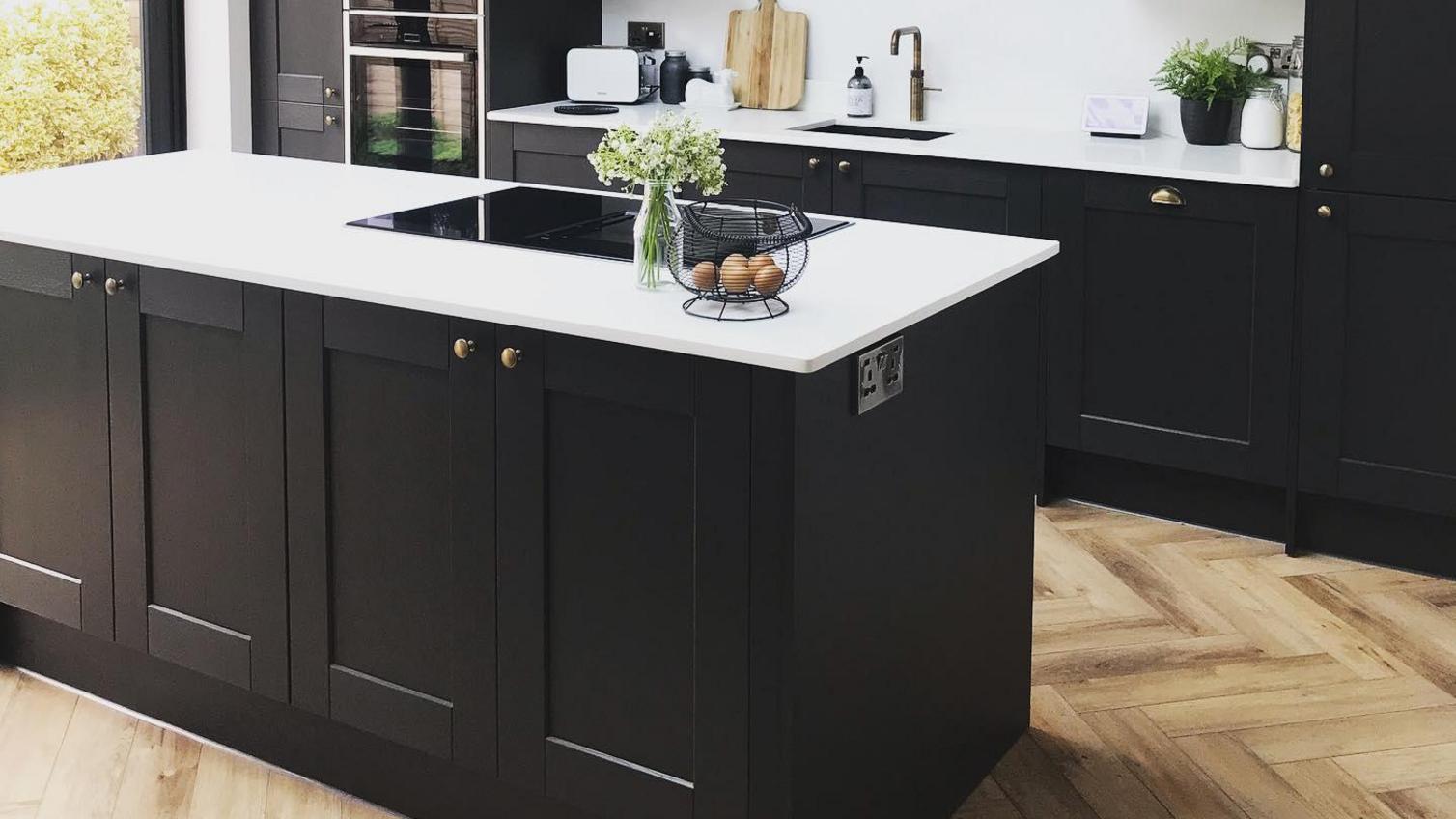 Black shaker kitchen with an island layout, with brass knob handles, chevron flooring and a white quartz worktop.