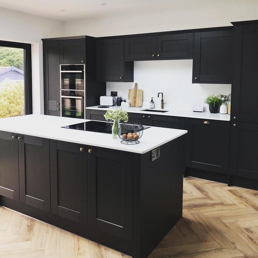 Black shaker kitchen with an island layout, with brass knob handles, chevron flooring and a white quartz worktop.