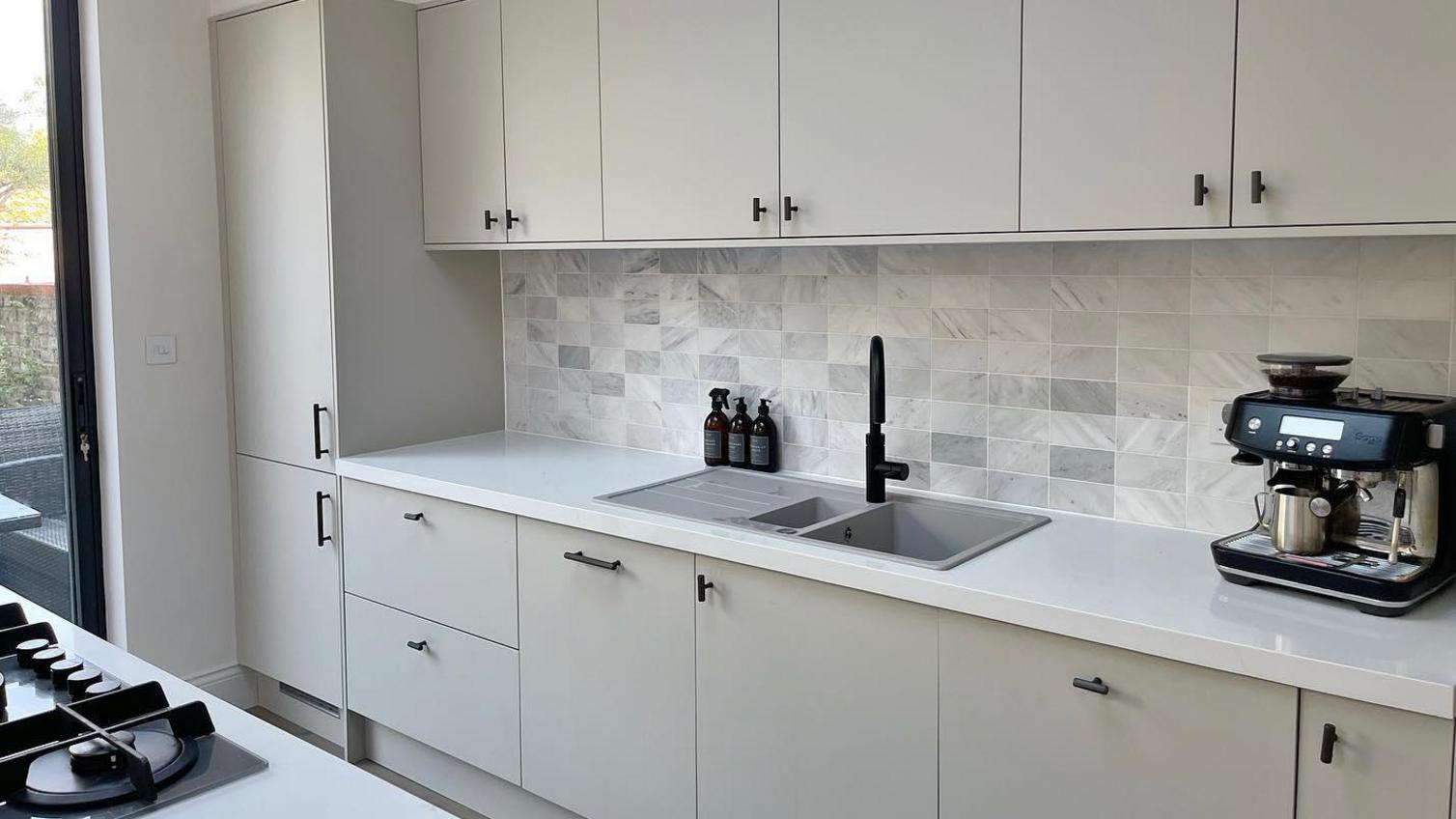 A simple cream kitchen idea using sandstone slab units. Features black handles, white worktops, and a grey, composite sink.